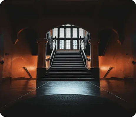 Dark, grand staircase leads to a bright arched window inside an old building.